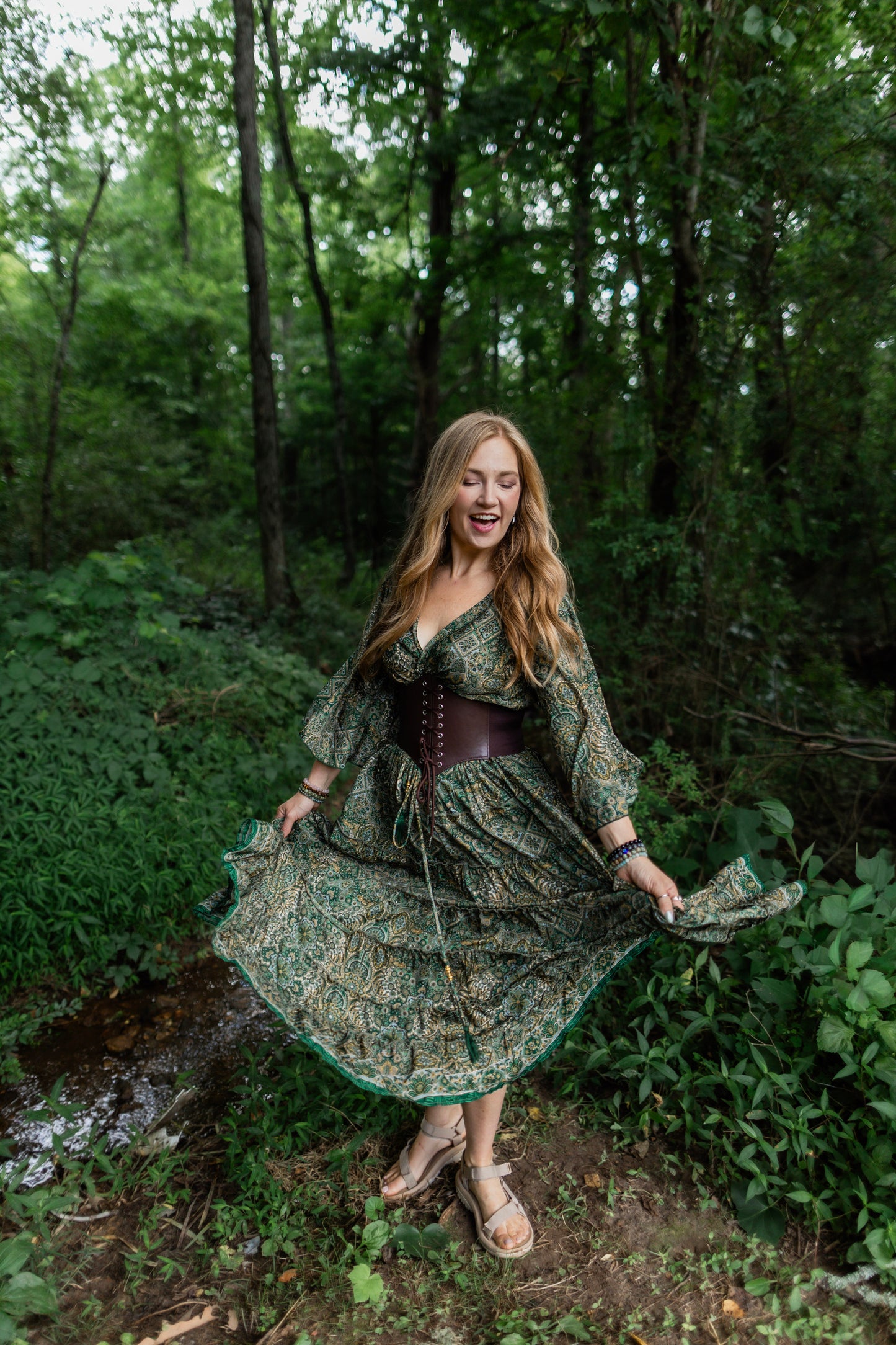 A woman stands in a wooded outdoor setting wearing a long-sleeve bohemian-style skirt set with a green and brown floral pattern, a dark brown corset-style bodice, and holding out the skirt to show its flow. She is wearing tan flat sandals.