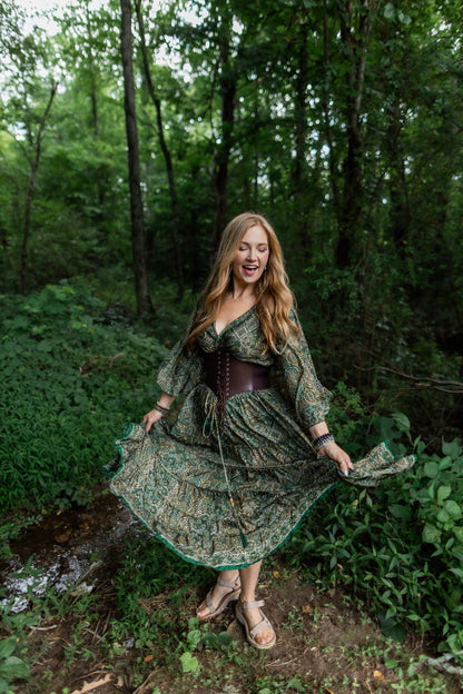 A woman stands in a wooded outdoor setting wearing a long-sleeve bohemian-style skirt set with a green and brown floral pattern, a dark brown corset-style bodice, and holding out the skirt to show its flow. She is wearing tan flat sandals.