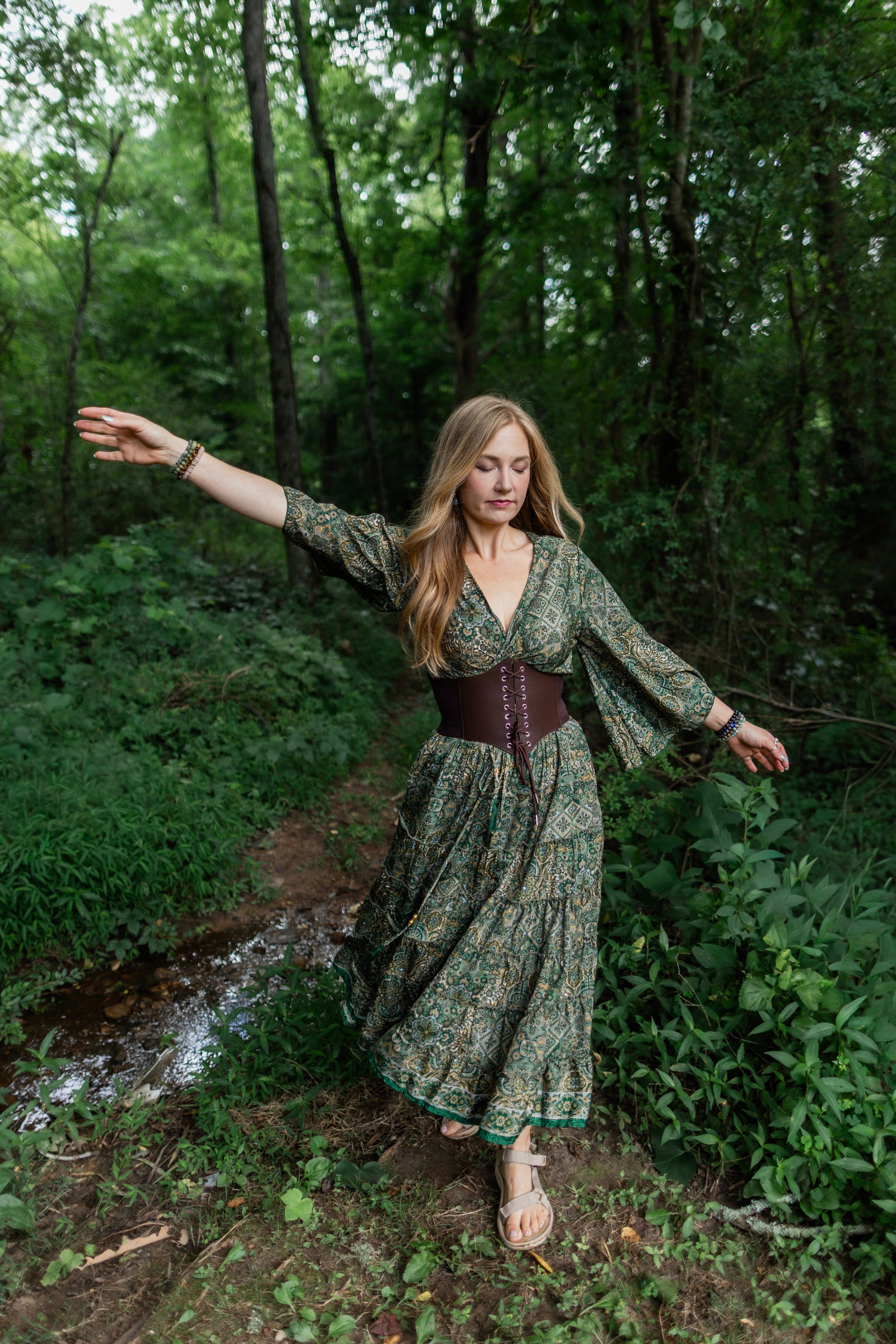A woman stands on a forest path wearing a long-sleeved, tiered green floral maxi skirt with a brown corset-style waist belt and bare feet.