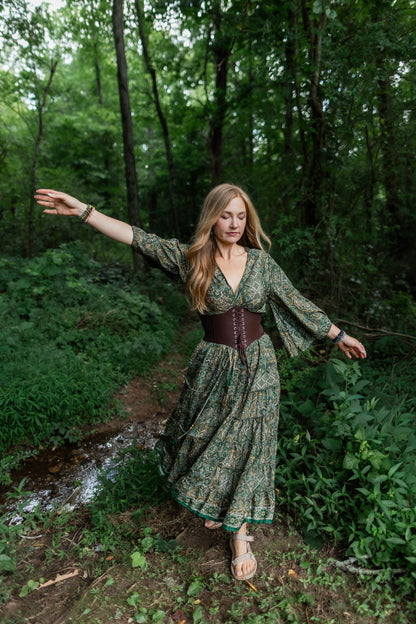 A woman stands on a forest path wearing a long-sleeved, tiered green floral maxi skirt with a brown corset-style waist belt and bare feet.