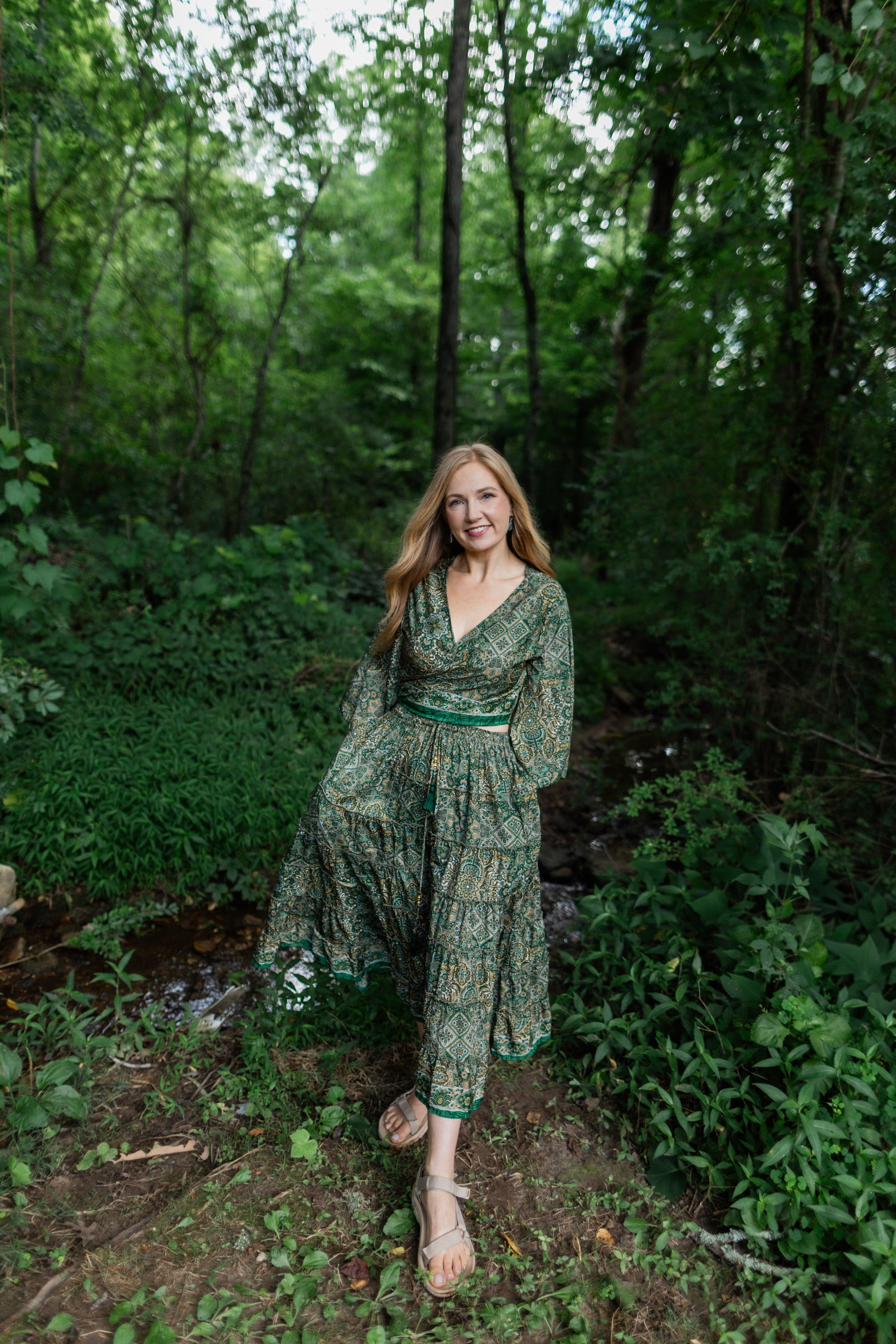 A woman wearing a green patterned bohemian maxi skirt set stands in a lush green forest clearing.