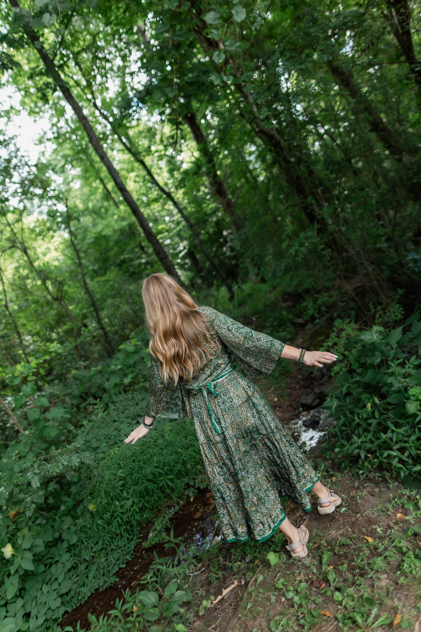 A woman with long blonde hair stands in a wooded forest clearing wearing a long-sleeved green patterned maxi set and beige sandals, viewed from behind as she looks down a dirt path.