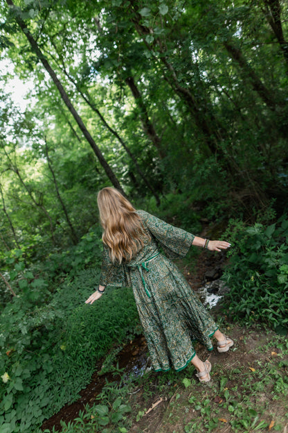 A woman with long blonde hair stands in a wooded forest clearing wearing a long-sleeved green patterned maxi set and beige sandals, viewed from behind as she looks down a dirt path.