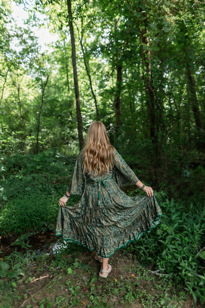 A woman stands in a wooded forest clearing wearing a long flowing green patterned skirt and top set, holding the skirt out to show its drape and print. The background is filled with tall trees and green foliage.