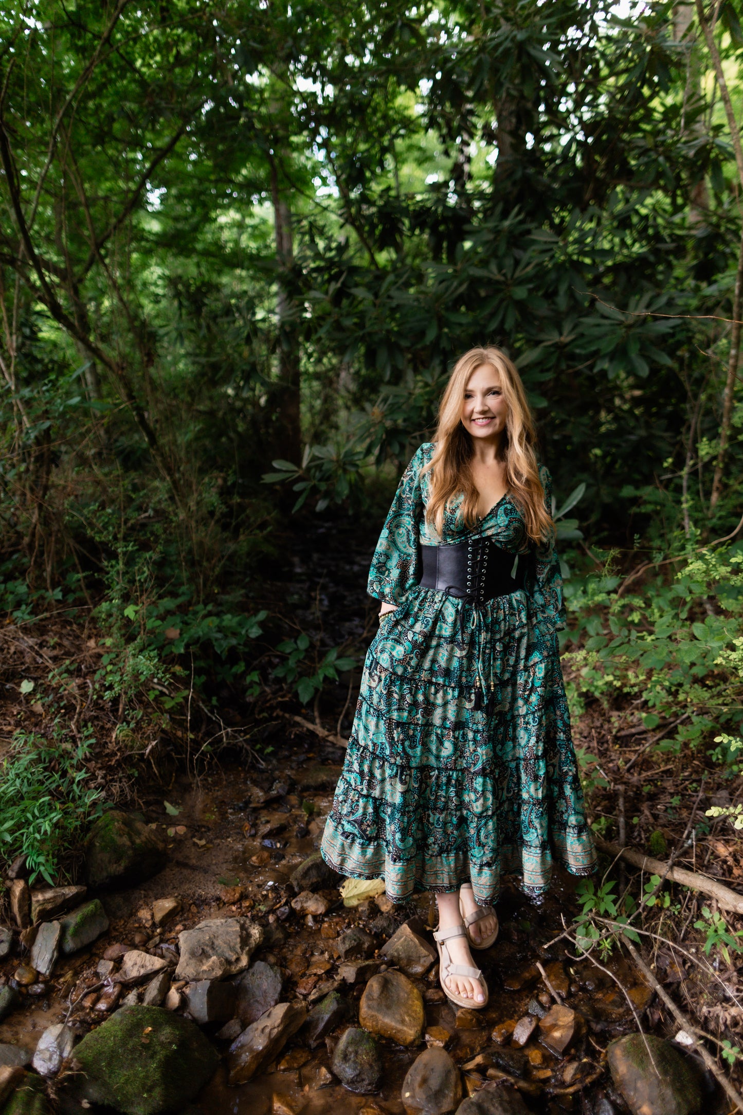 A woman stands in a rocky forest stream wearing a long green floral maxi dress with a black corset-style waist and sandals.