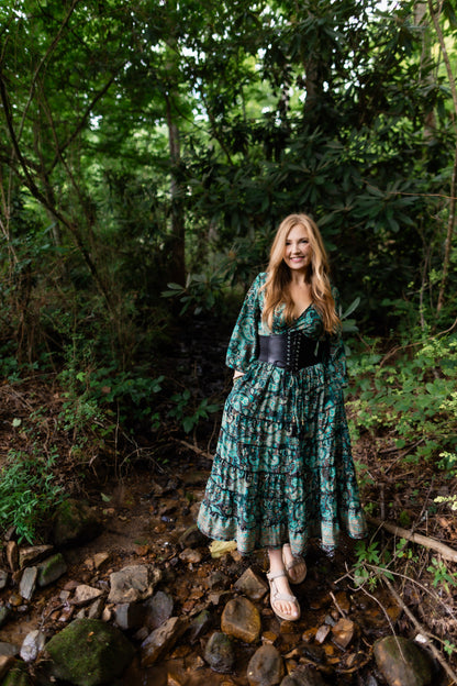 A woman stands in a rocky forest stream wearing a long green floral maxi dress with a black corset-style waist and sandals.