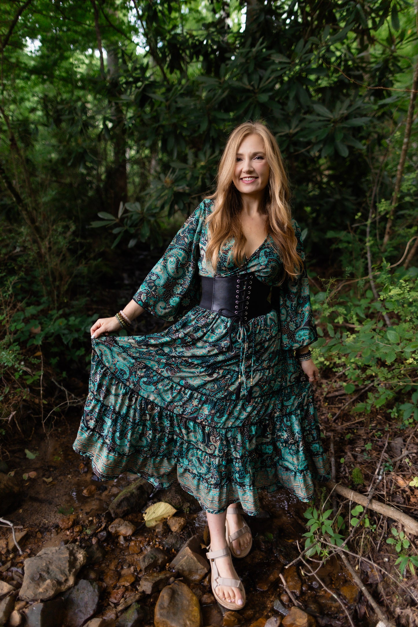 A woman stands in a rocky forest stream wearing a long teal and black patterned maxi dress with a black corset-style waist, holding out the skirt to show its flow. The background is a dense green woodland setting.