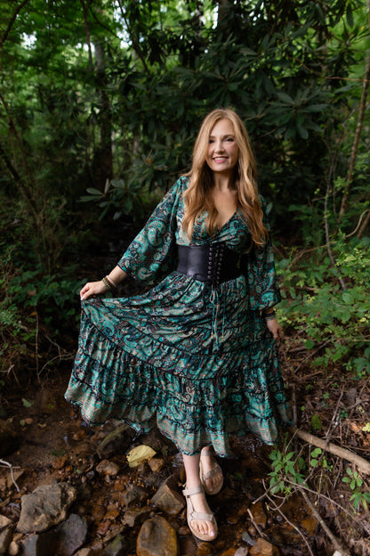 A woman stands in a rocky forest stream wearing a long teal and black patterned maxi dress with a black corset-style waist, holding out the skirt to show its flow. The background is a dense green woodland setting.