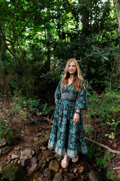 A woman stands in a rocky forest stream wearing a long teal and green floral print maxi dress with three-quarter sleeves and a tie waist.