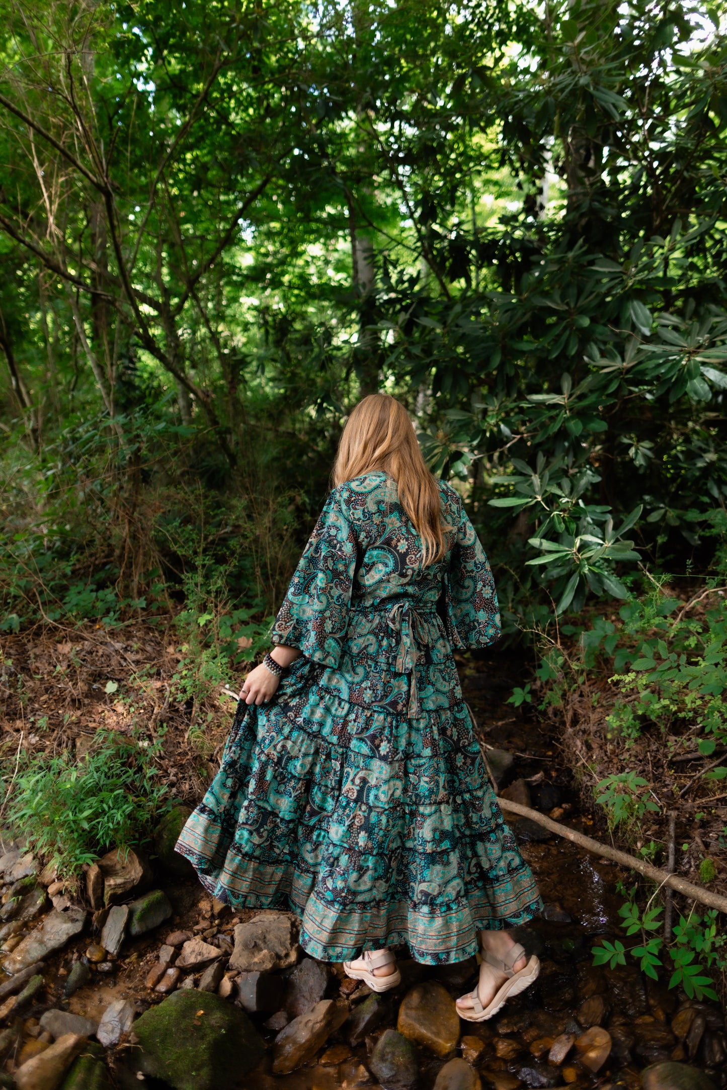 A woman viewed from behind wearing a long teal and green floral print maxi dress while walking on rocks in a wooded stream.
