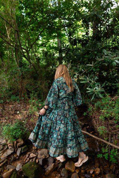 A woman viewed from behind wearing a long teal and green floral print maxi dress while walking on rocks in a wooded stream.