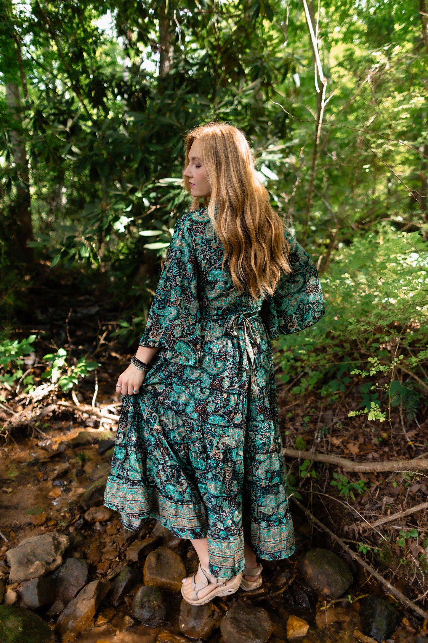 A woman with long blonde hair stands on rocks in a forest stream, wearing a long-sleeved teal and black patterned maxi dress with a tie waist.