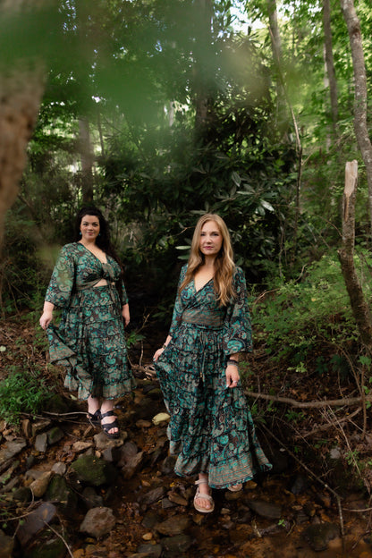 Two women wearing matching green floral maxi sets stand on a rocky stream in a dense forest. The dresses feature a dark green base with a lighter green and teal abstract floral pattern, and the women are wearing sandals.